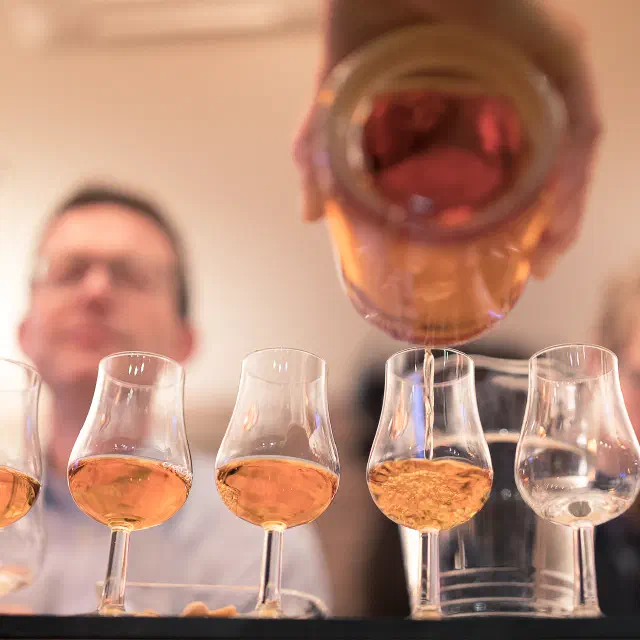 Eine Reihe Nosing-Gläser auf dem Tresen, in eins wird Whisky aus der Flasche eingeschenkt, dahinter zwei Gäste unscharf