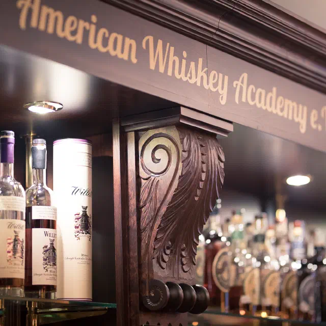 Flaschen Willett Bourbon im Regal unter einem geschnitzten Holzbalken mit dem Schriftzug American Whiskey Academy e.V.