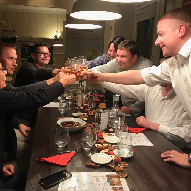 Gäste an einer gedeckten Tafel heben lachend ihre Whiskygläser zum Anstoßen, im Hintergrund eine Karte der schottischen Brennereien